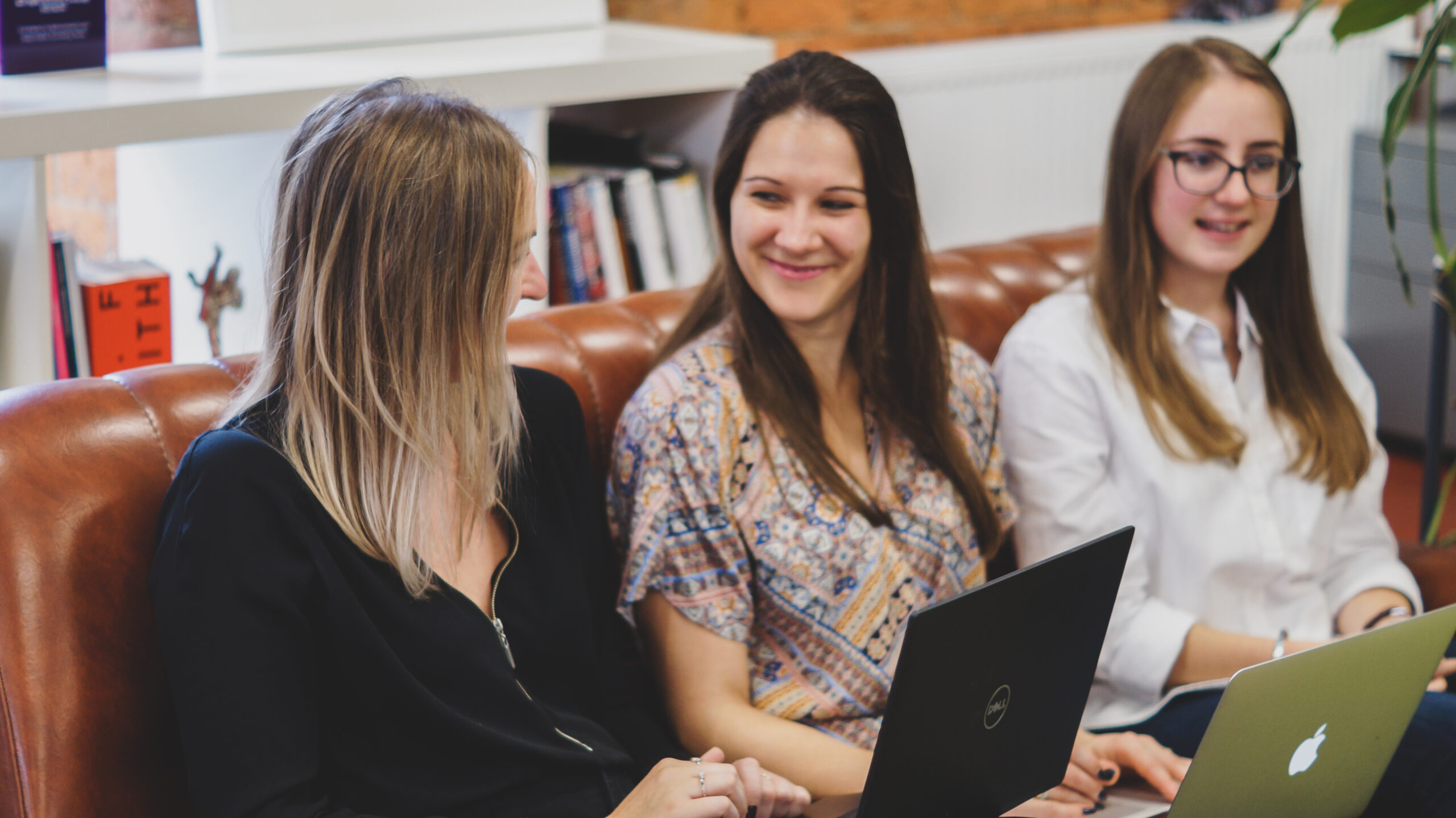 Three women sitting on a sofa with computers at their laps. Talking and smiling.